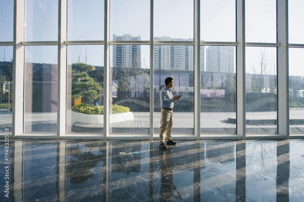 Fototapeta premium Man standing in front of glass curtain wall while looking at his smartphone