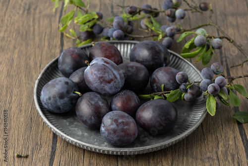 Bild auf Leinwand Retro plate with whole and sliced fresh ripe garden plum fruits and wild organic blackthorn branches with ripe berries on wooden table background