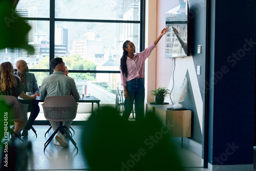 Young businesswoman in smart casual pointing at screen while presenting to team in bright office
