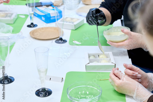 Professional confectioner hands in gloves making handmade white chocolate truffles at master class workshop. Group of people learning to decorate desserts in kitchen studio with glasses and tools.