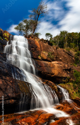 Diagonal cascade waterfall flowing over warm colored rocky cliffs beneath a solitary tree under a dramatic blue sky in a tropical mountain landscape.