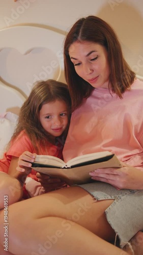 Mother and daughter share a moment as they read a book together in a cozy bedroom during the evening,vertical video