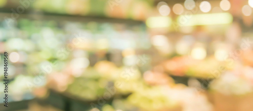 Blurred image of a grocery store interior showcasing various produce items. Colorful fruits and vegetables are displayed in baskets on shelves with warm lighting.