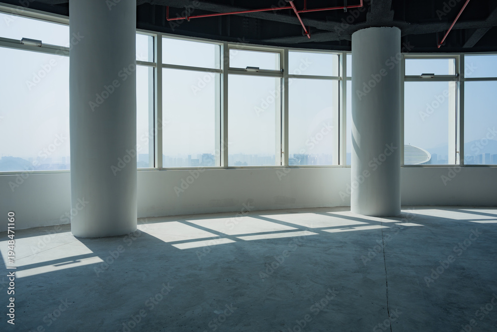 Fototapeta premium Spacious empty office interior with floor-to-ceiling windows, white columns, and natural light casting geometric shadows