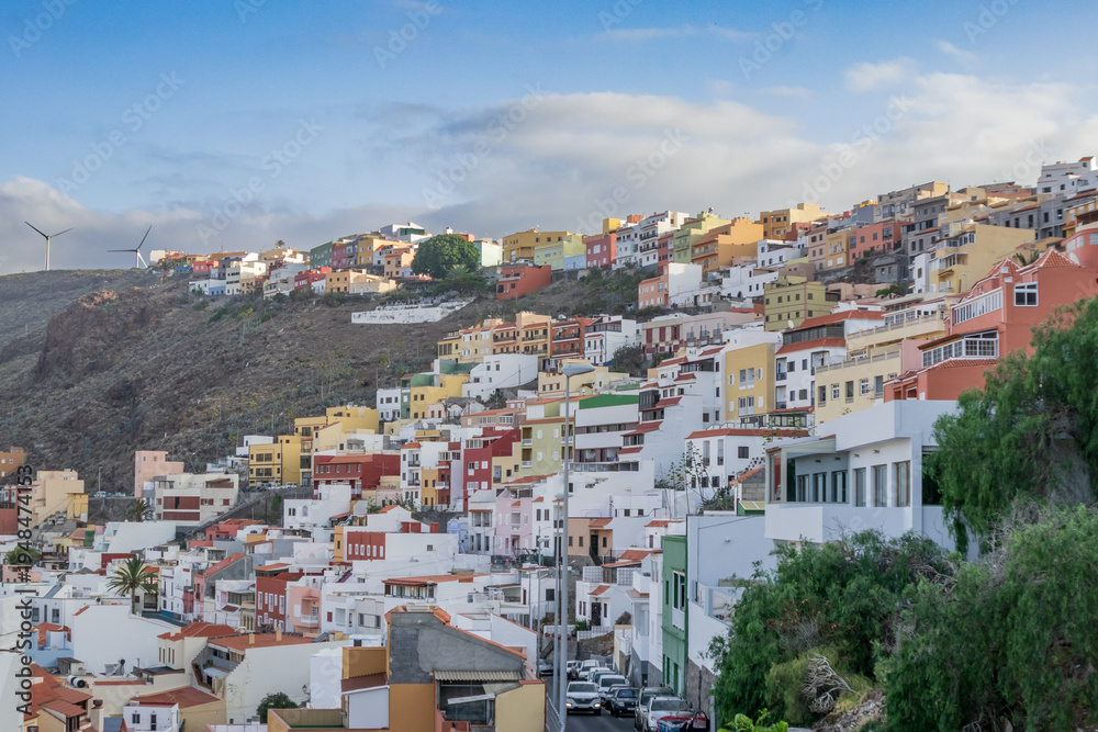 Fototapeta premium San Sebastián de la Gomera: pintorescas casas de colores escalonadas sobre la ladera de la montaña. Paisaje urbano canario con molinos eólicos, cielo suave y arquitectura típica de la isla.