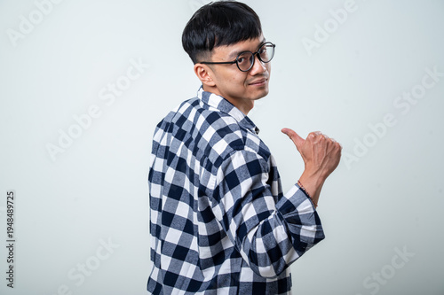 Young Asian man in round glasses and a plaid shirt looks back over his shoulder while giving a thumbs-up against a light gray studio background
