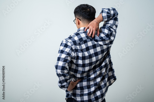 Young Asian man seen from behind wearing a plaid shirt, scratching the back of his neck while standing against a light gray studio background