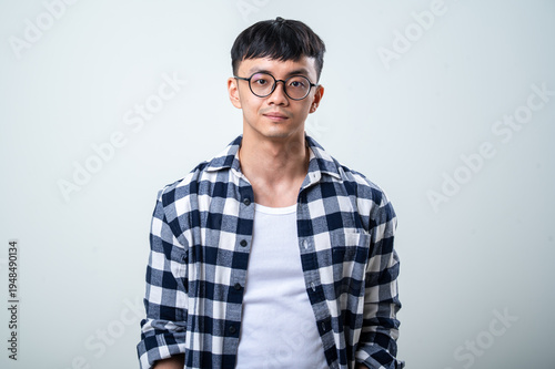 Young Asian man wearing round glasses, a plaid shirt, and white tank top stands against a light gray background, looking at the camera with a calm, casual expression