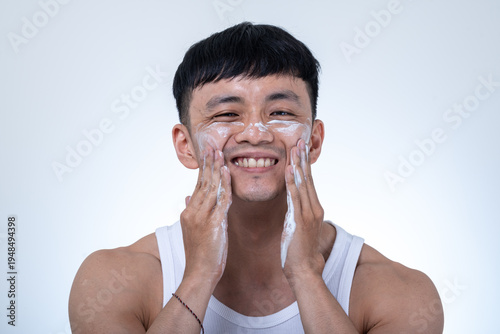 Young Asian man in a white tank top applies white facial cream to both cheeks with his hands against a light gray studio background