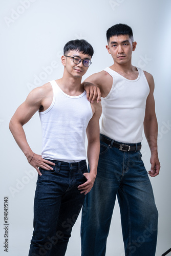 Two young Asian men in white tank tops and jeans pose confidently in a studio, standing together against a light gray background