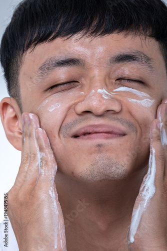 Young Asian man in a white tank top applies white facial cream to both cheeks with his hands against a light gray studio background