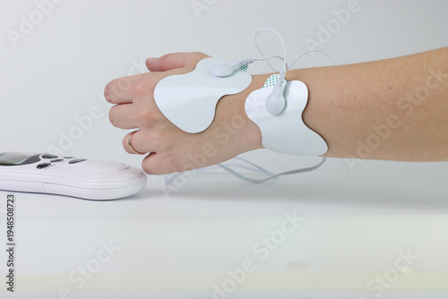 A person's wrist and hand are shown with TENS unit electrode pads attached for pain relief therapy