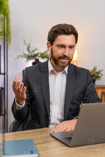 Middle-aged businessman at home office joins video conference on laptop wearing wireless earphones. Freelancer guy at table smiles listens and takes notes in note book during meeting.