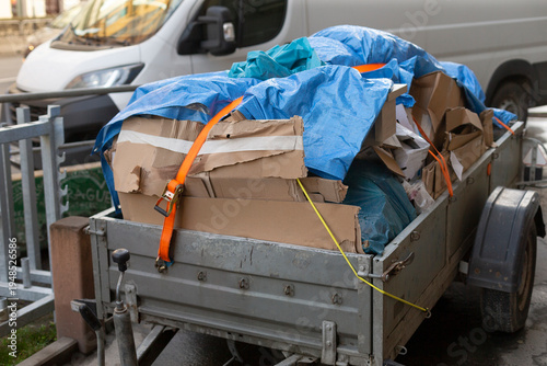 Overloaded trailer packed with cardboard, blue tarp secured by orange ratchet strap, metal utility trailer hitched