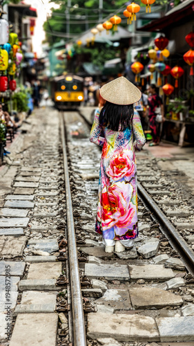 Woman walking along Hanoi train street with colorful Vietnamese lanterns and vintage steam locomotive approaching between narrow houses in Vietnam.