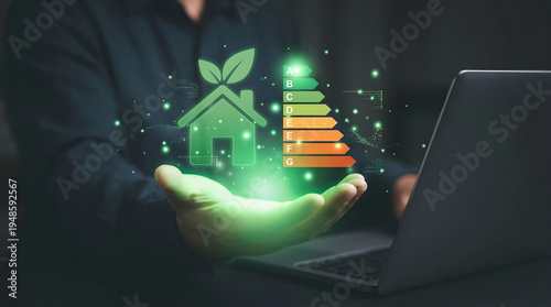 A glowing green hand presents a digital icon of a sustainable house with leaves and an energy efficiency rating scale A to G alongside a laptop in a d