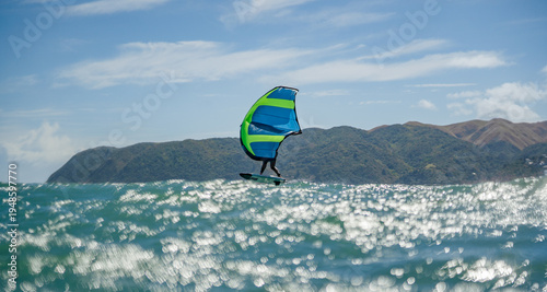 A man wing foils at sea using a hand held inflatable bright green and blue wing, riding a hydrofoil surfboard.  Turquoise sea, cloudy blue sky. 