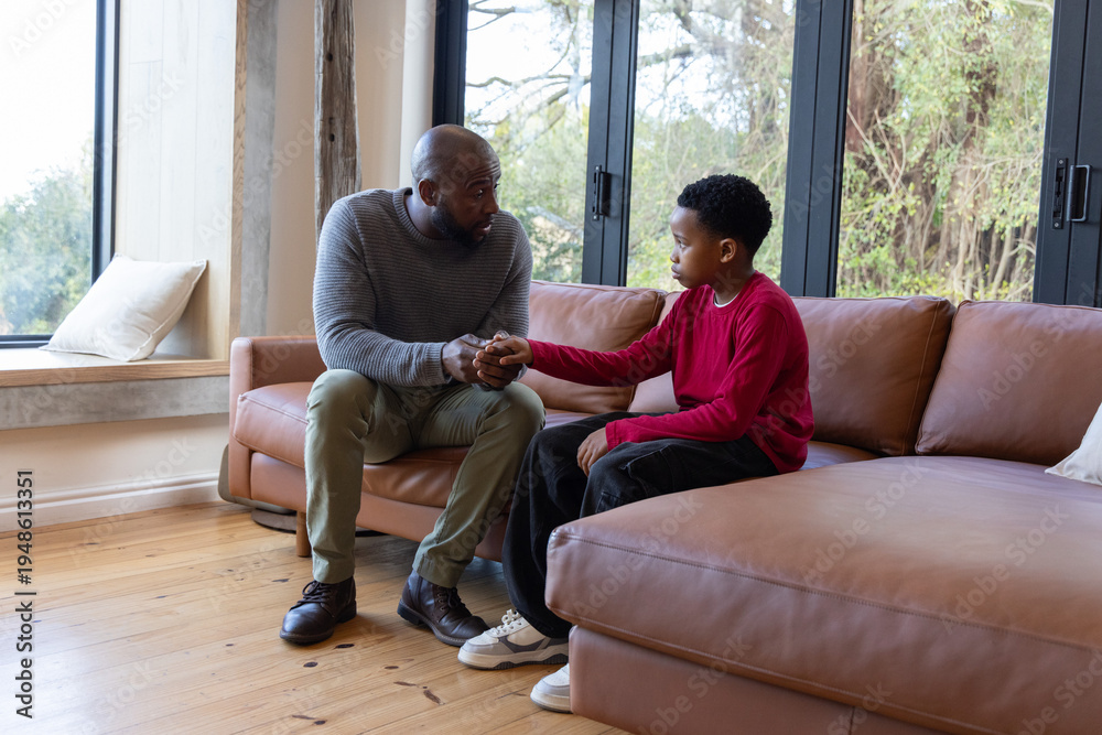 Obraz premium African American father and son sitting on tan leather sectional in living room, holding hands