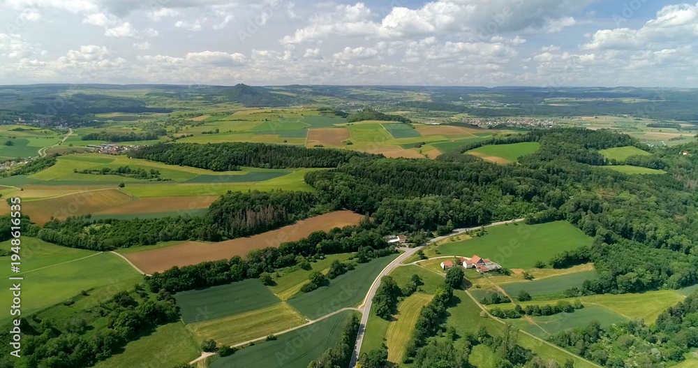 Fototapeta premium Green fields and hills stretch across the countryside with a winding road and small houses below in summer light