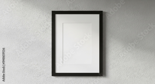 A minimalist black wooden photo frame with a white mat board hanging on a rough textured white plaster wall, creating a modern interior gallery look.