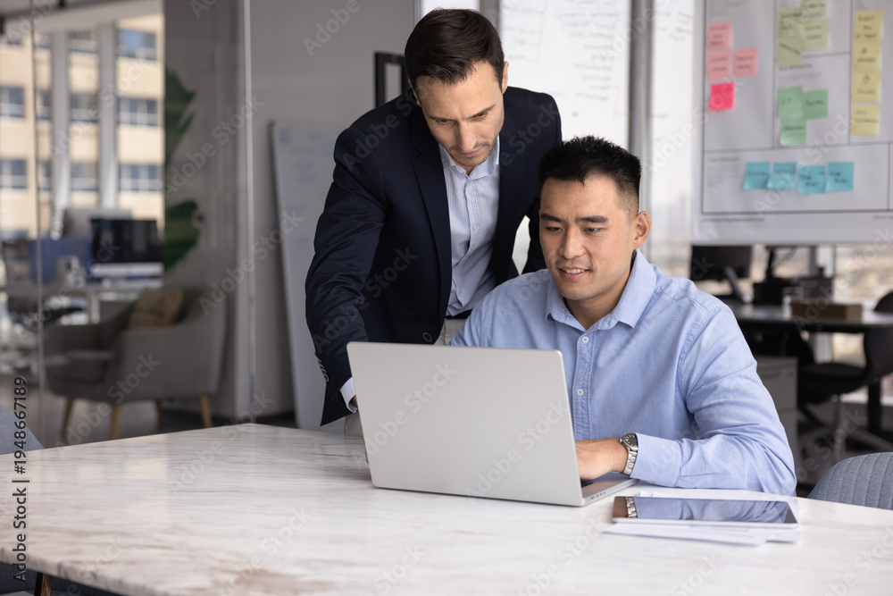 Obraz premium Workplace guidance and assistance. Experienced mid adult businessman in formal suit stand close to seated male intern trainee review laptop screen give consultation expertise feedback help with work