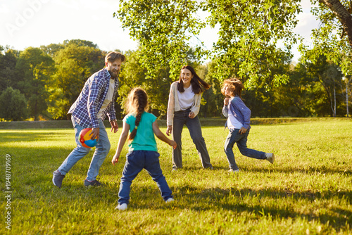 Fotografie Cheerful parents and two children playing ball together on green lawn in park on sunny day