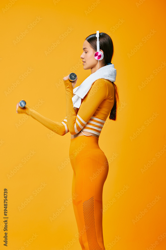 Obraz premium Female athlete in bright orange activewear lifts dumbbells in a bold studio, wearing headphones and a towel around her neck, showcasing strength, focus, and energetic fitness training in a vivid