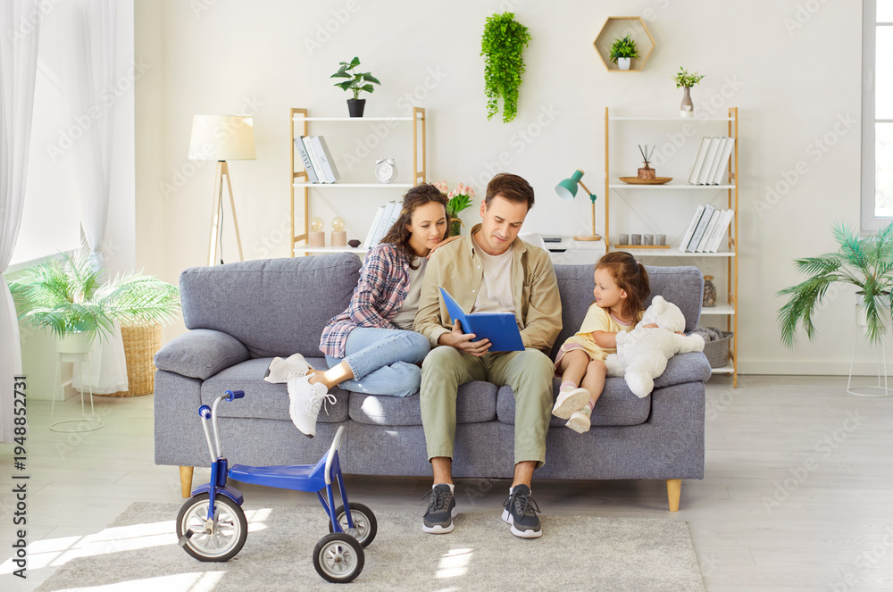 Fototapeta premium Parents reading fairy tale book with daughter at home together. Father sitting between little kid and woman on sofa, holding book to read children literature to listening family in living room