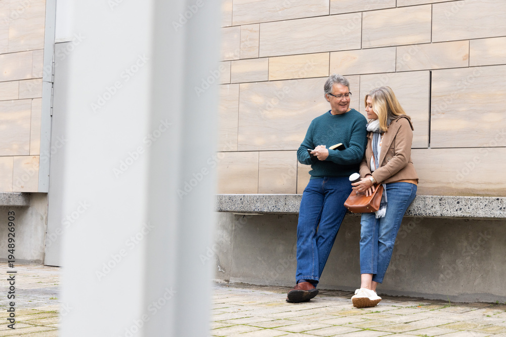 Fototapeta premium Senior couple leaning against concrete ledge in plaza holding book and coffee cup, copy space