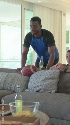Vertical video: Leaning man in blue tee reacting to TV at home, getting on couch and sipping bottle