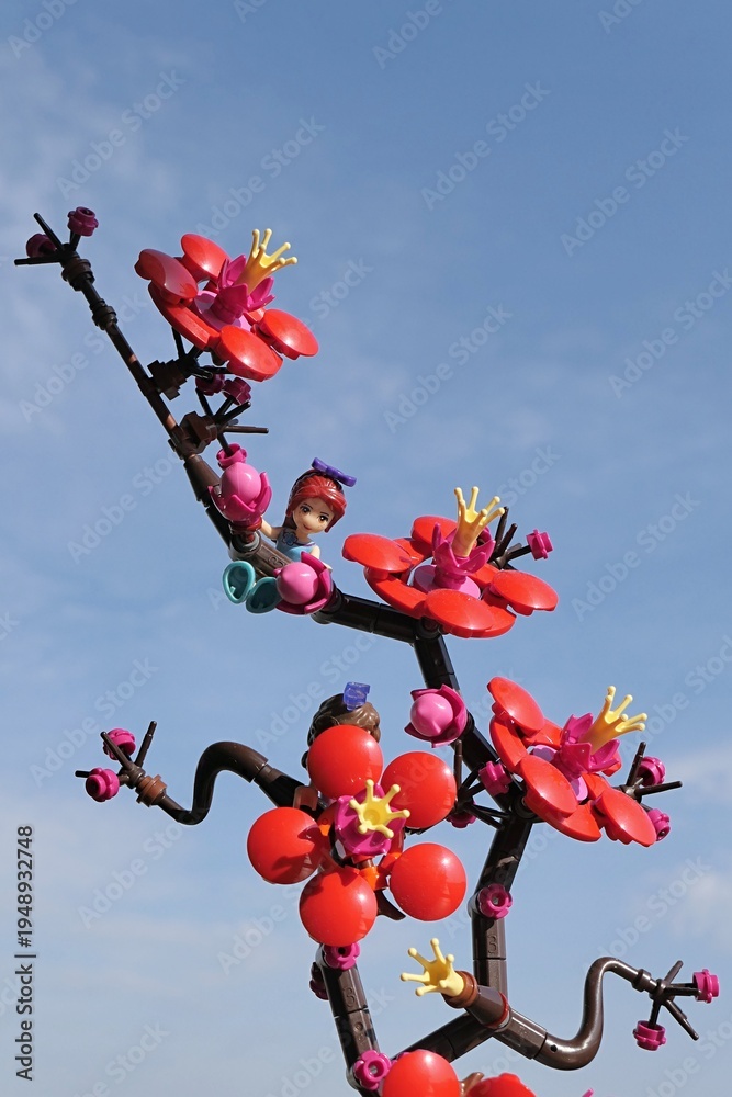 Fototapeta premium LEGO Friends figure of Mia hanged on LEGO Botanicals Plum Blossom branch, blue skies with some scarce clouds in background, spring morning sunshine. 