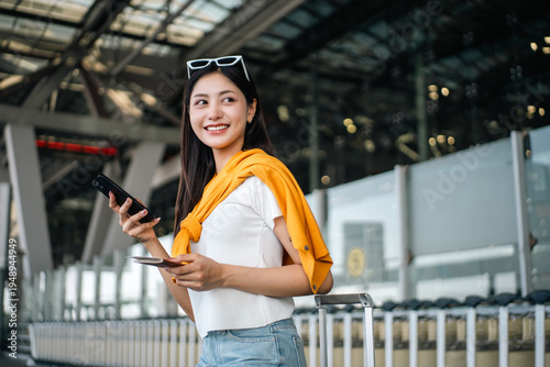 Young asian woman traveler using smartphone for checking flight information check the routes trip while waiting with her luggage at the airport terminal. Traveller woman passport and baggage luggage