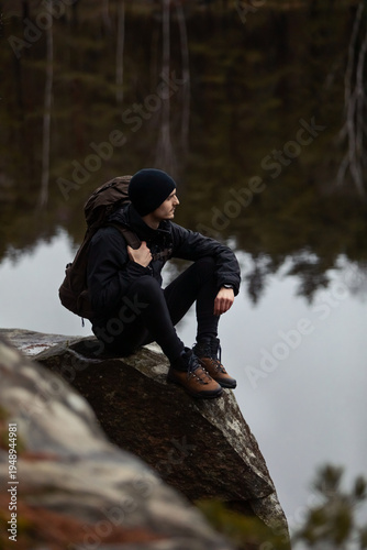 Photos Hiker in black outdoor gear sitting on a large rock by a calm forest lake reflecting trees