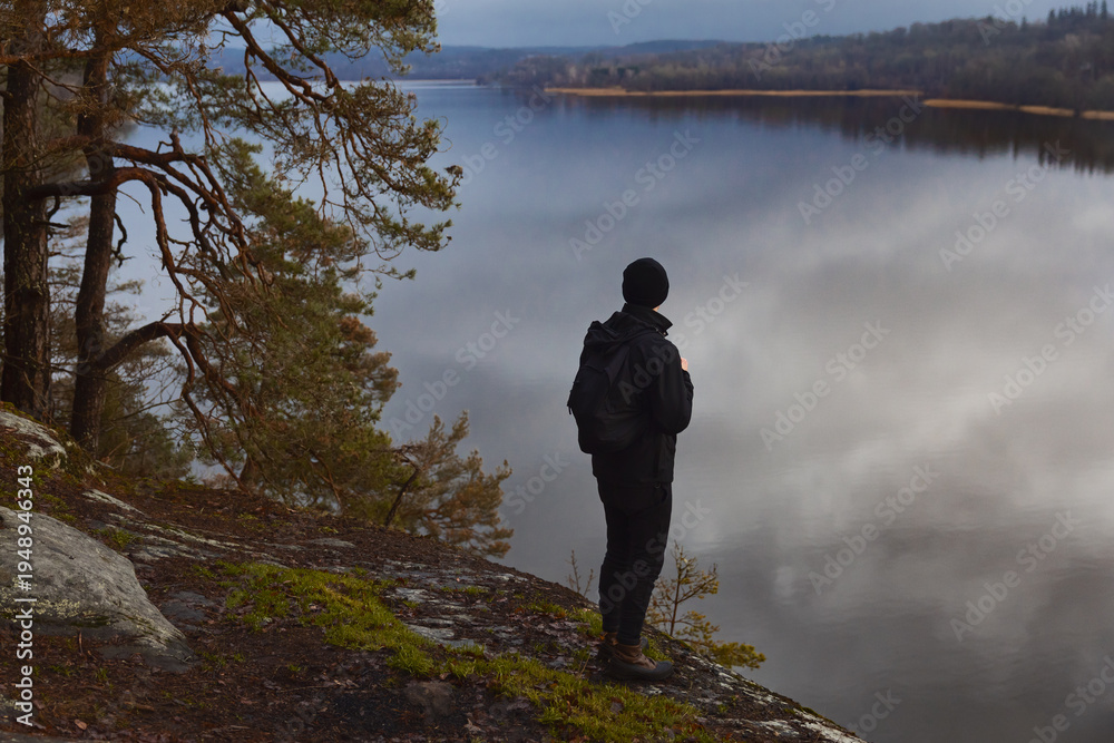 Fototapeta premium A man in dark hiking gear standing on a rocky cliff, looking out over a calm, misty lake at dawn. A peaceful, atmospheric scene of solitude and Scandinavian nature during a moody autumn morning.