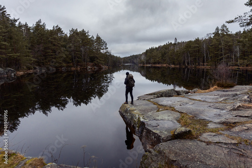 Canvas Print Hiker with a backpack standing on a rocky cliff edge, overlooking a calm, mirror-like lake surrounded by a dense pine forest under a moody, overcast sky