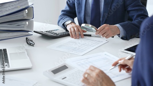 Male financial analysts with female colleague examining documents, using magnifying glass and calculator at workplace in office. Audit and taxes in business