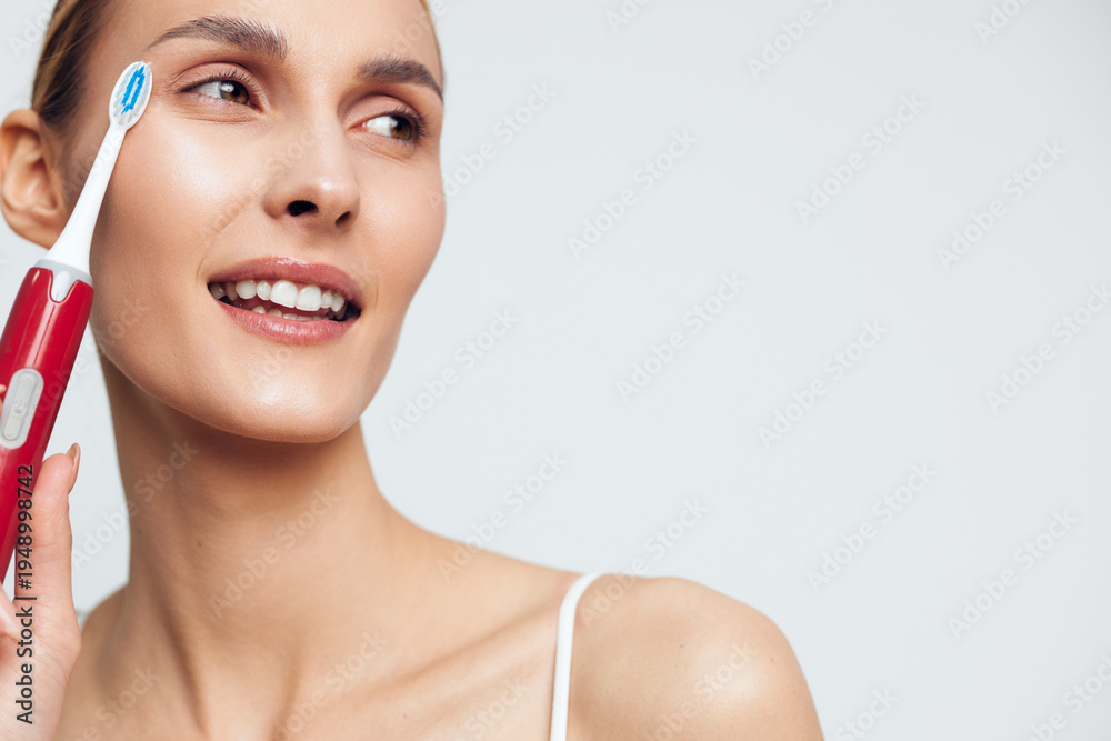 Obraz premium Young woman holding electric toothbrush near face smiling with clean skin looking aside isolated on white background personal hygiene dental care concept.