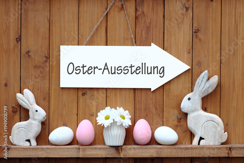 Holzwand mit zwei weißen Hasenfiguren, Ostereiern und einer Vase mit weißen Blumen auf einem Schild steht der Text Osterausstellung.