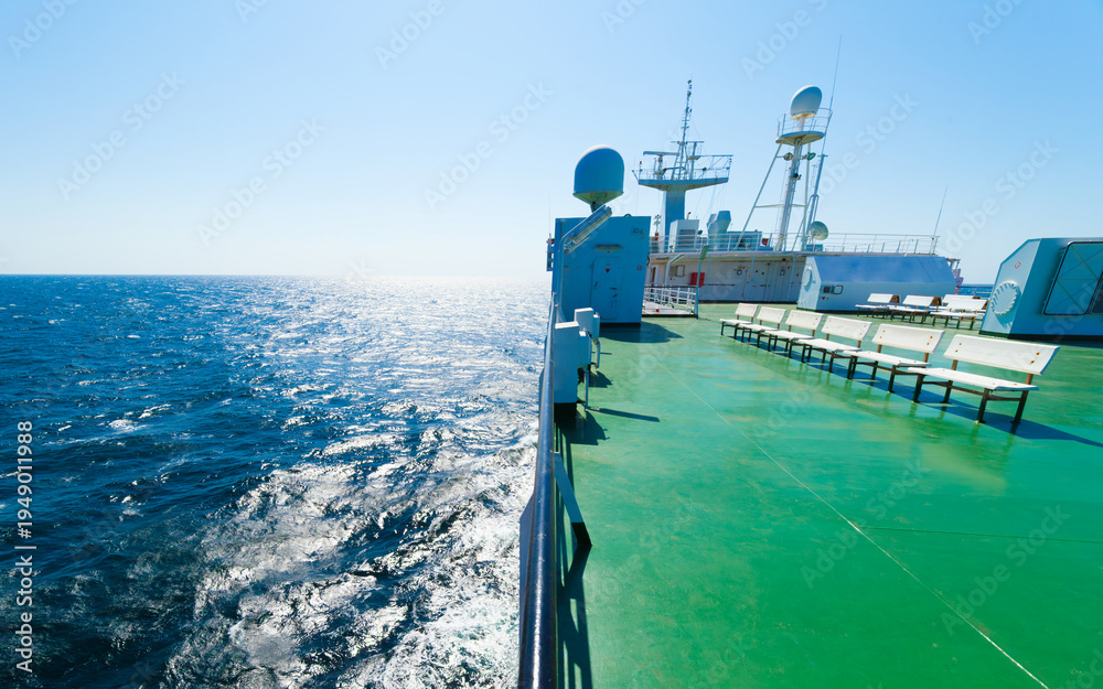Naklejka premium Empty passenger deck of large ferry ship sailing on deep blue sea under clear sky. White benches stand on green floor of ferry near navigation equipment and radar domes