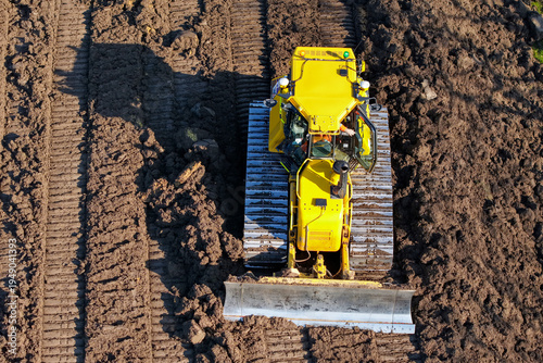 Yellow bulldozer works on field soil during sunny day in rural area with clear blue sky