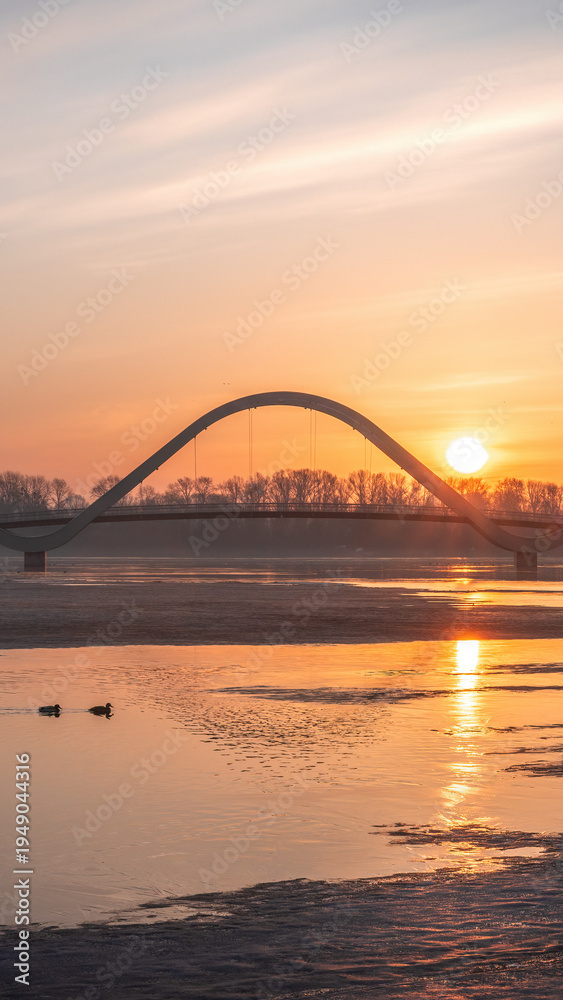 Obraz premium Golden sunrise over the modern pedestrian bridge across the Dnipro River in Kyiv, Ukraine. Calm water, winter riverbanks and silhouettes create a peaceful morning city landscape.