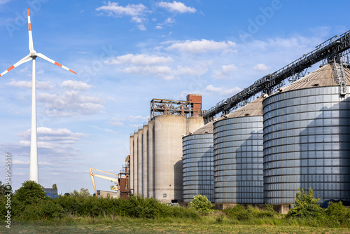 Large agricultural grain silo conveyor system industrial storage facility area wind turbine blue sky. Rural industry infrastructure combine crop storage logistic operation renewable energy production