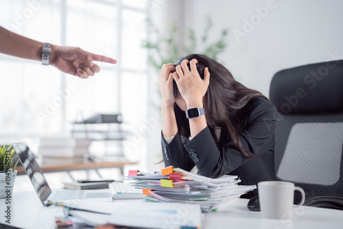 Frustrated employee facing pressure and blame from a pointing boss, feeling overwhelmed by workload and stress while working at her office desk surrounded by documents