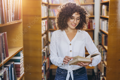 Woman with curly hair and glasses standing in a library, smiling and reading a book between bookshelves