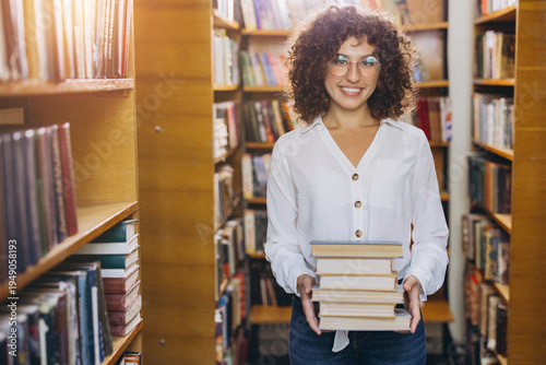 Canvastavla Happy female student in library, smiling while carrying a stack of books, repres
