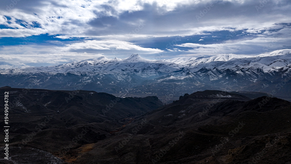 Fototapeta premium Aerial view of majestic snow-capped mountain peaks and dark rugged valleys under a dramatic cloudy sky