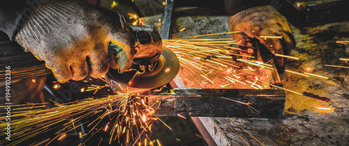 Worker hands wearing protective gloves operating an angle grinder, creating a shower of bright orange sparks while cutting or shaping metal in an industrial workshop.