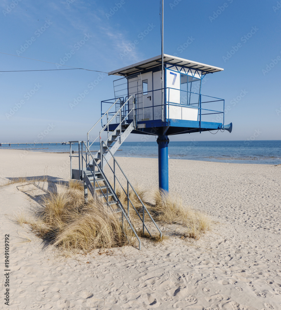 Fototapeta premium A modern blue and white lifeguard rescue station stands prominently on a wide, sandy beach under a clear blue sky