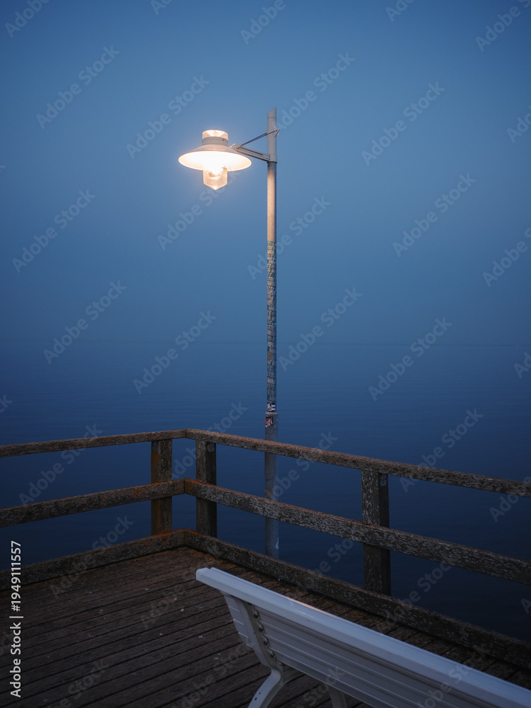 Fototapeta premium A glowing street lamp stands at the end of a wooden pier in dense blue fog, with a white bench in the foreground and calm water merging into the misty horizon