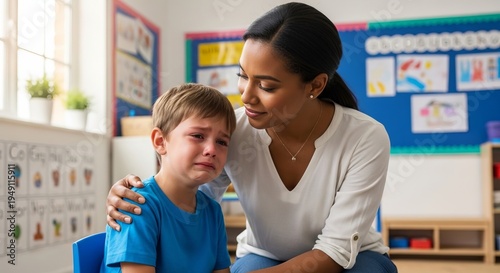 Caring teacher comforts crying schoolboy in bright classroom, offering emotional support during early childhood learning time